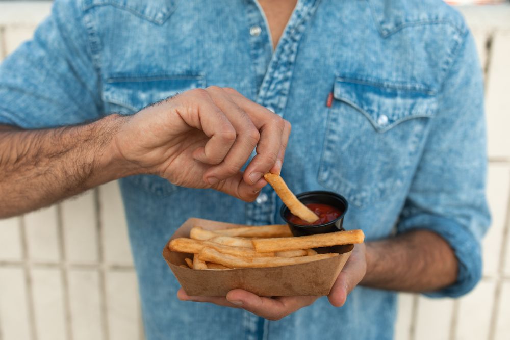 Cropped image of man denim shirt eating fries with ketchup