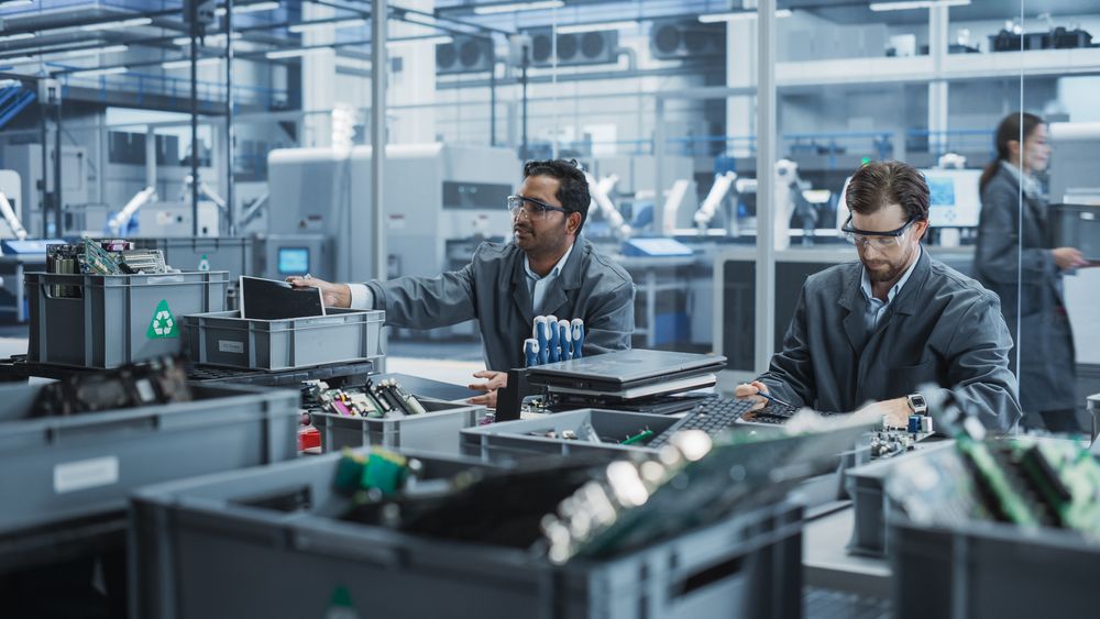 Indian And White Male Workers Taking Apart Laptops To Recycle For Production Of High-Tech Devices At Automated Electronics Factory With Robotic Arms.