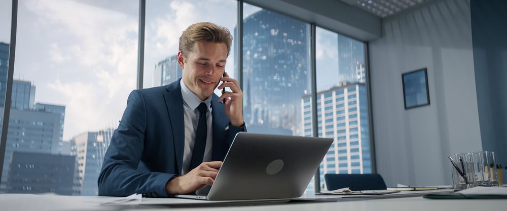 Confident Businessman in a Suit Sitting at a Desk in Office, Using Laptop Computer and Picking Up A Call
