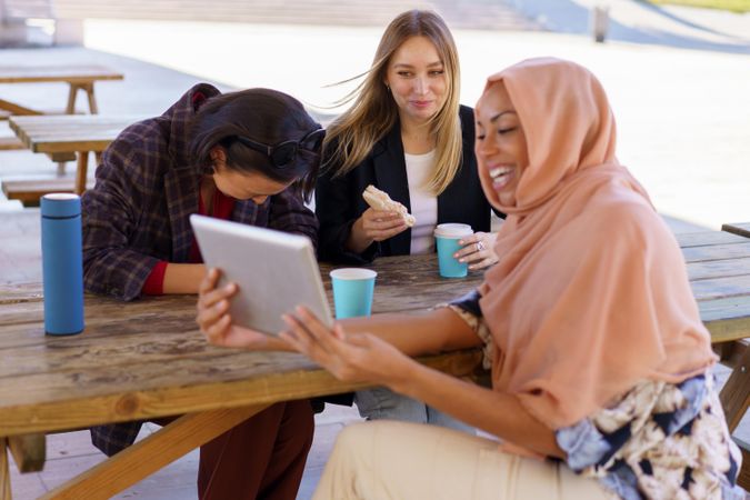 Three women laughing on park bench with insulated mugs and digital tablet