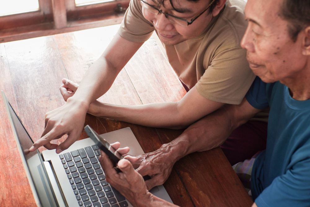 Happy adult son teaching older father using laptop and smartphone