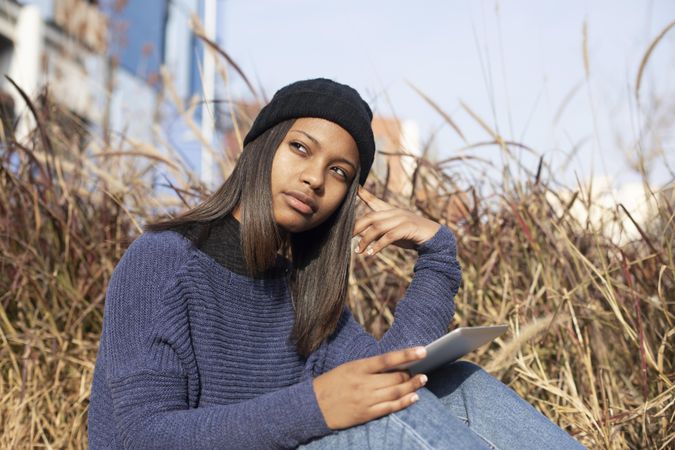 Curious female in hat and sweater sitting outside with tablet, copy space