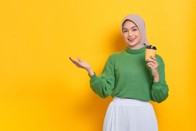 Woman in headscarf holding to go coffee cup and gesturing to the copy space