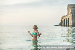 Woman in bikini standing in the calm sea with her eyes closed and hands raised 0JERdb