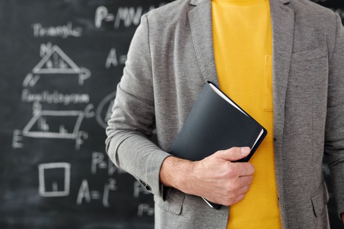 Cropped image of math teacher holding a book standing against chalkboard