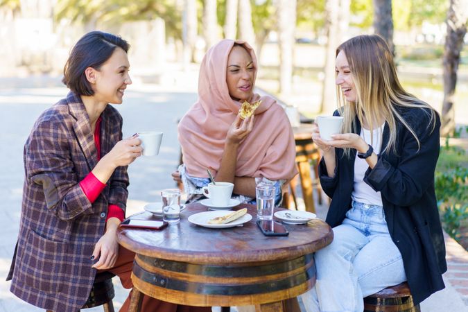 Three women talking and laughing over coffee and baked goods on sunny outside table
