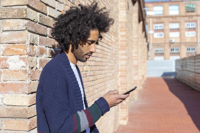 Side view of Black male looking down at his smartphone while leaning on a brick wall outdoors on sunny day