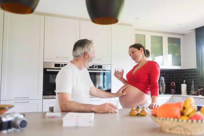 Pregnant woman in kitchen with glass of water as her husband touches her stomach