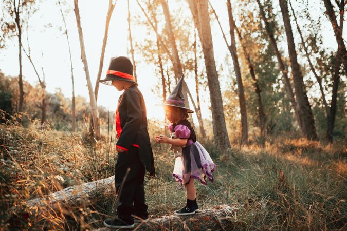 Young brother and sister dressed as witch and skeleton playing on log in the forest