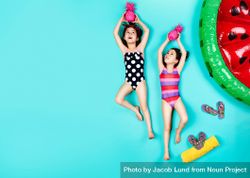 Twin girls in swimwear lying down on floor holding colored pineapple with slippers 5RpENb