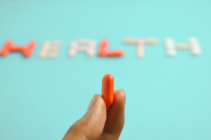 Hand holding pill in front of "HEALTH" spelled on blue background