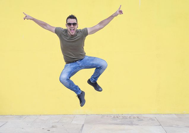 Smiling male jumping in front of yellow wall with outstretched arms