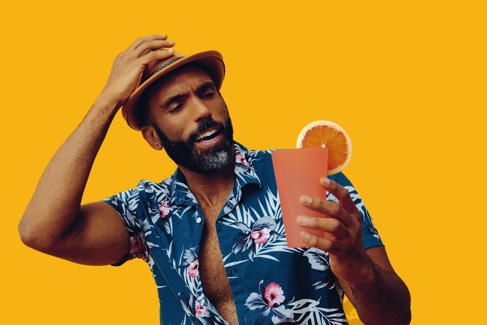 Male wearing colorful shirt and looking at cocktail with yellow background