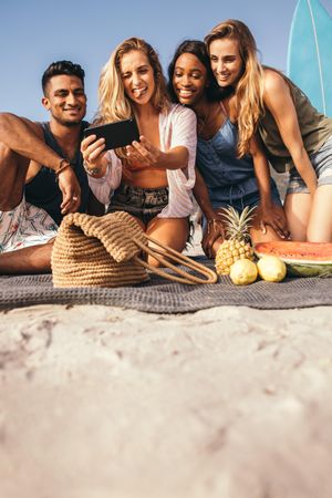 Ground level shot of group of friends sitting on beach having fun taking selfie on mobile phone
