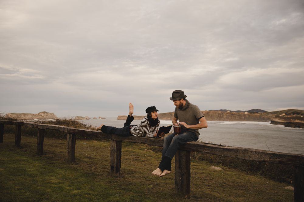 Man plying on drum near woman with notebook on bench on shore