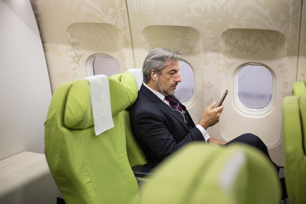 Male sitting in window seat watching phone screen with ear buds in flight