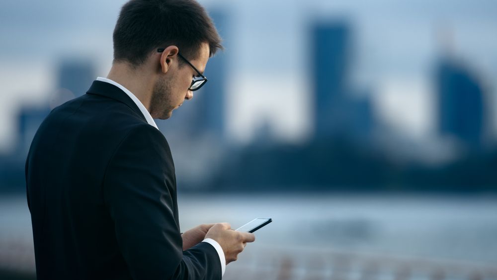 Young Businessman Uses Smartphone while Standing on the Street with Big City Skyscrapers View.