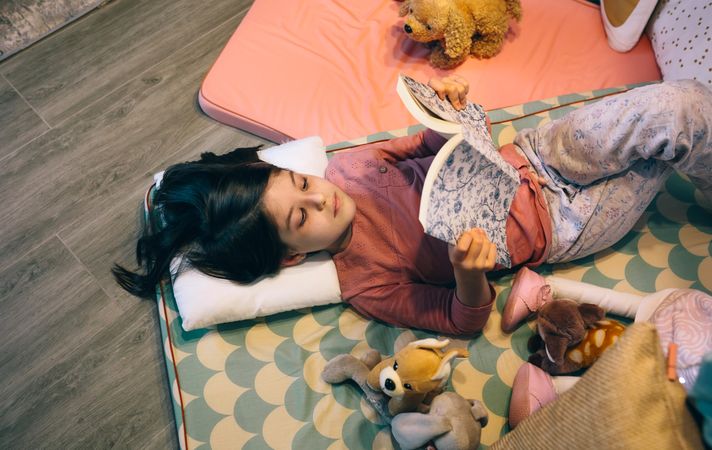Top view of serene little girl looking a book while resting over cozy sleeping mats in living room