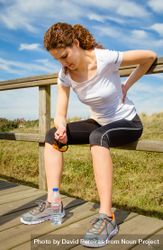 Woman in athletic gear holding back while seated on wooden path 48VRX0