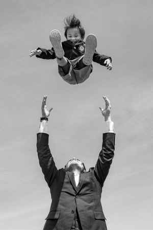Grayscale photo of a father wearing suit throwing his son to the air