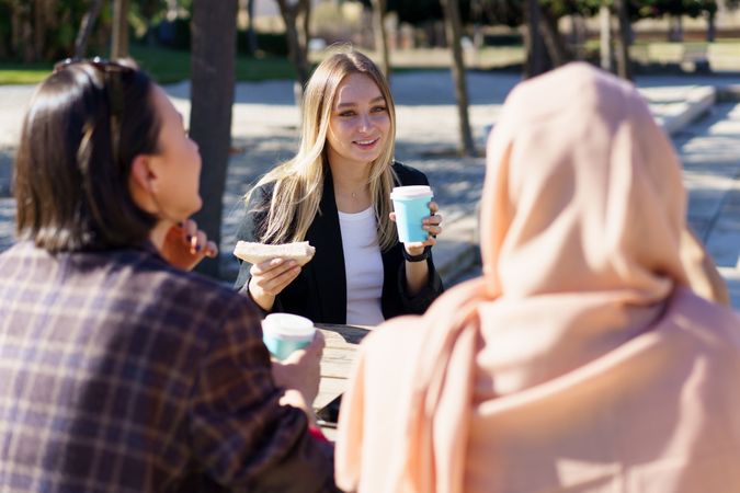 Blonde woman holding sandwich and coffee cup chatting with her friends outside