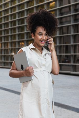 Smiling Black pregnant woman talking on smartphone and carrying a laptop