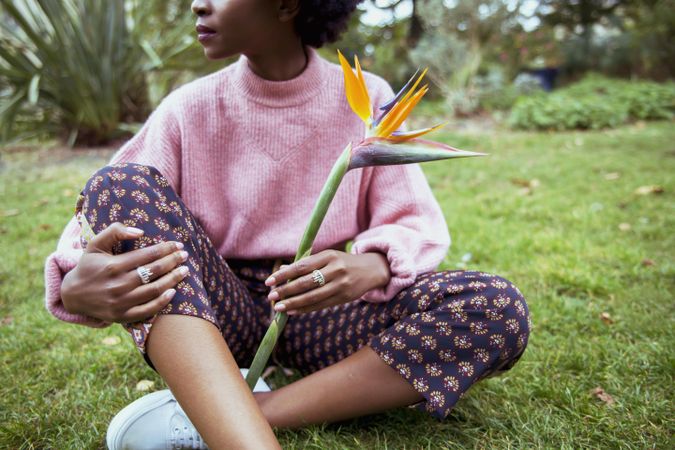 Woman in pink sweater holding a flower and sitting on grass field