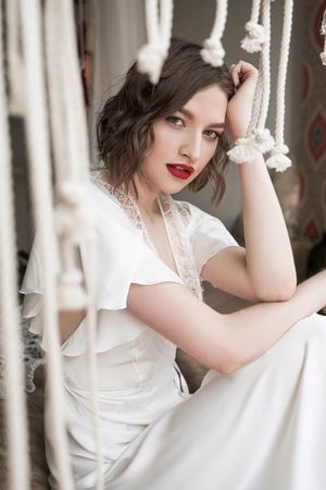 woman with red lips in white dress looking at camera while sitting on floor beside sofa