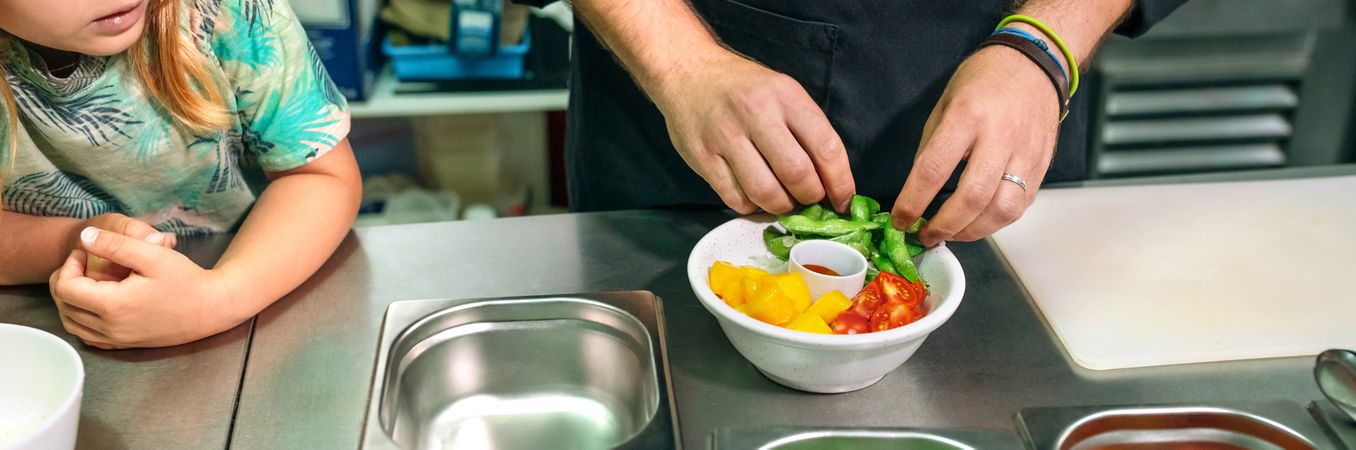 Unrecognizable chef preparing vegan poke bowl while little girl watching him in professional kitchen