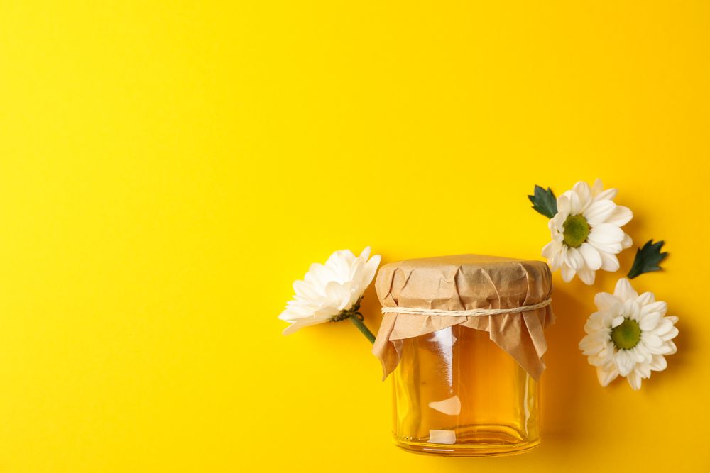 Glass jar with honey and chamomile on yellow background, space for text