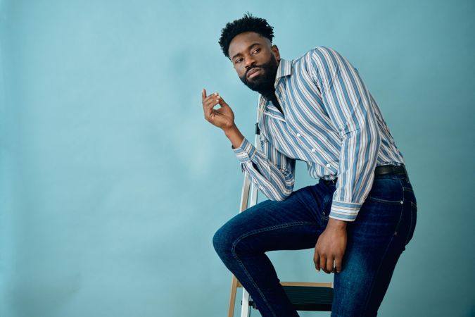 Black male model in striped blue shirt and jeans posing casually in blue studio on step ladder