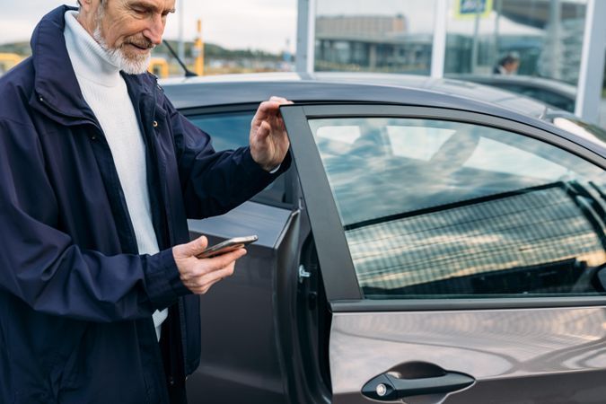 Man with grey beard opening a door of his car while looking at his phone
