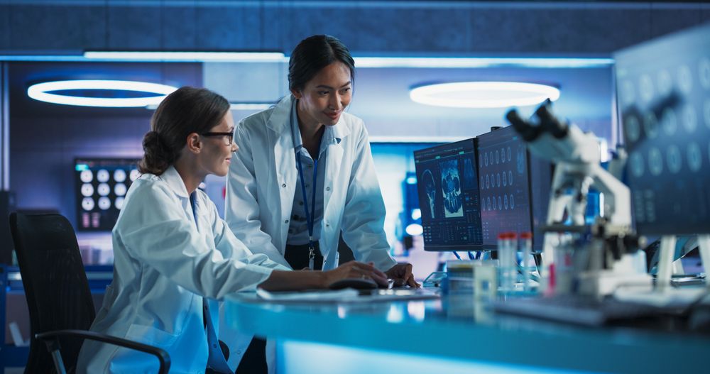 Asian And White Female Doctors Talking In Medical Research Center And Using Desktop Computer To Analyze Brain CT Scans.