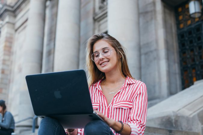 Chic woman sitting on outdoor steps in European city smiling at her laptop