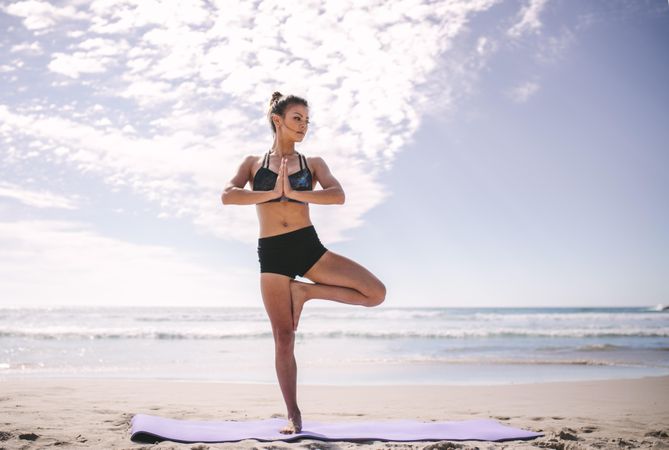 Woman practicing yoga with hands together and standing with one leg on the beach