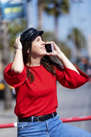 Side view of young woman wearing red blouse sitting on a metallic fence while using a mobile phone outdoors in the street on a bright day