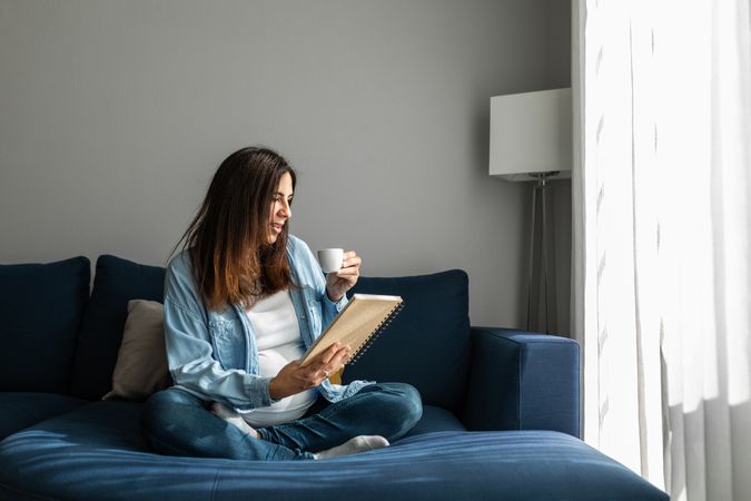Cheerful pregnant woman drinking beverage on sofa at home