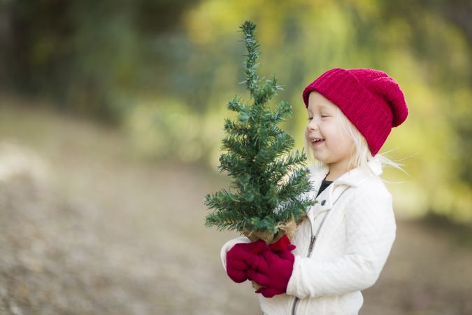 Baby Girl In Red Mittens and Cap Holding Small Christmas Tree