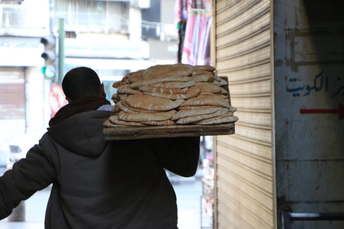 Back view of man holding a a large of wooden tray of bread walking in an alley