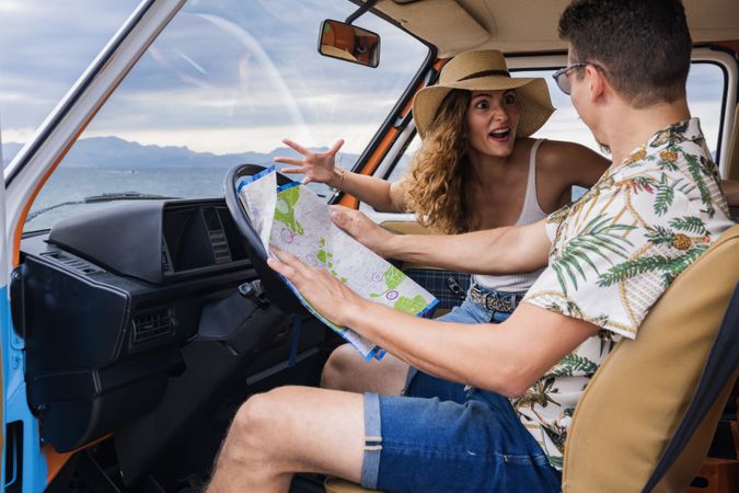 Touristic man exploring map with help of long haired woman in hat in front seat of car
