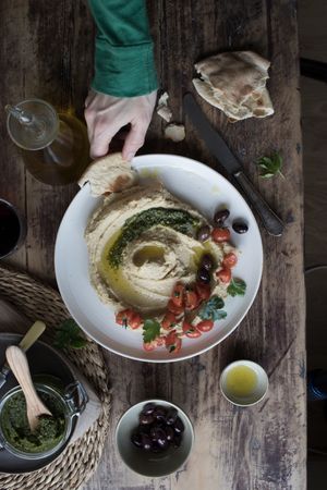 Crop hand dipping flatbread into hummus pesto with tomatoes and beans