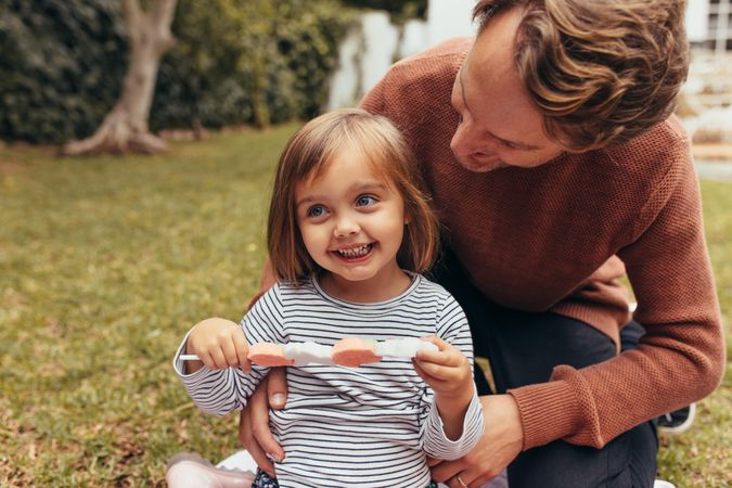 Smiling girl sitting with her father outdoors holding large candy