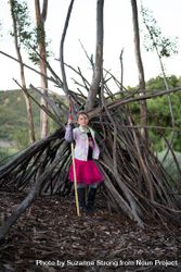 Confident young girl standing in front of tree fort holding a walking stick 5kRp30