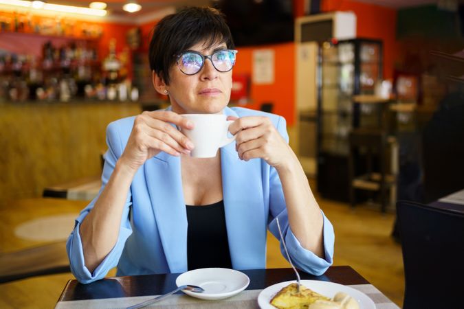 Positive female in trendy blue jacket and eyeglasses sitting at table with cup of hot drink and lost in thought
