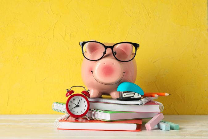 Happy piggy bank with glasses, and school accessories on wooden table against color background, space for text. Finance, saving money