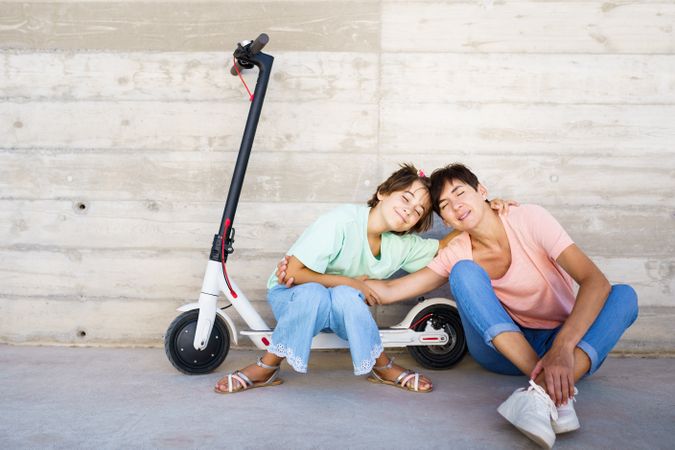 Woman and preteen girl sitting on the ground outside posing with a scooter