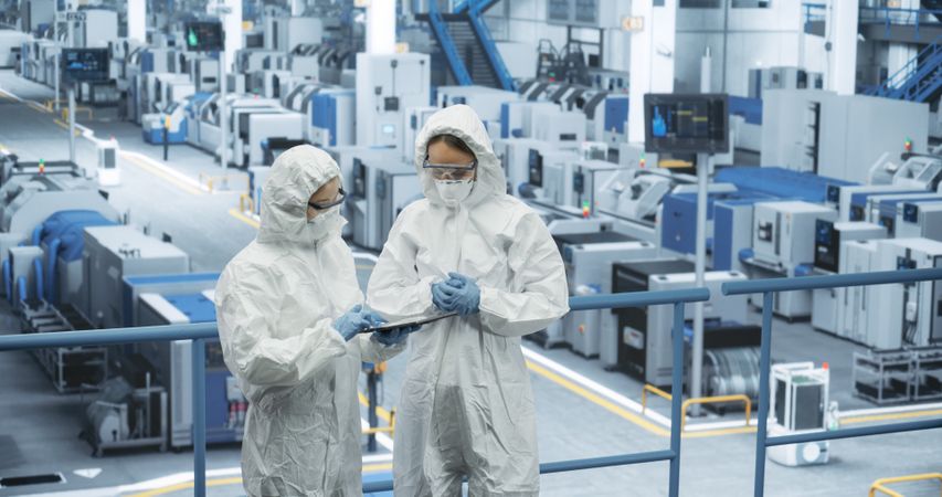 Female Workers Wearing Full-Body Polypropylene Coveralls at a Manufacturing Plant. Specialists Using Tablet Computer, Discussing Automated Work Process. Employees Protected From Hazardous Materials