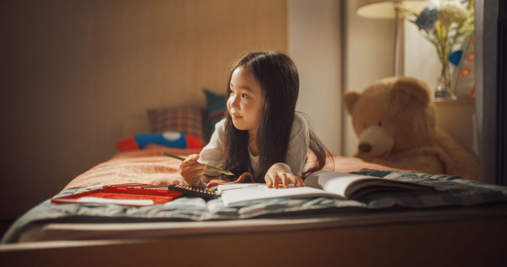 Portrait of a Little South Korean Girl Drawing with Colored Pencils in a Notebook While Lying in Bed in Children's Bedroom at Home.