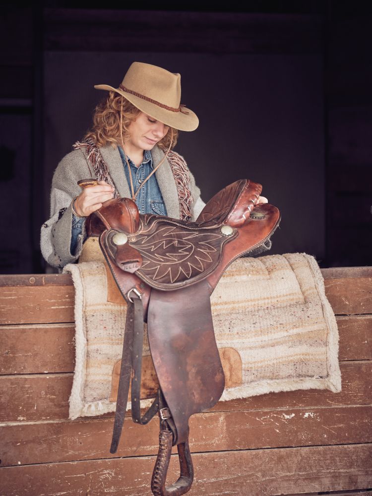 Woman in cowgirl hat placing saddle on carpet in stall