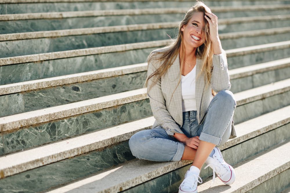 Woman in blazer sitting on outdoor steps on sunny day with hand to head
